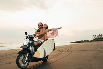 happy boyfriend and girlfriend riding scooter with surfboard on beach in bali, indonesia