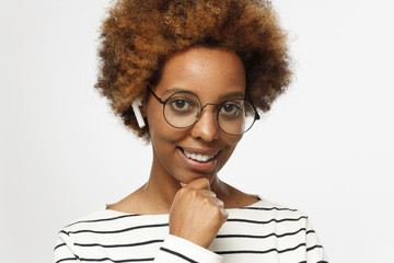 Smiling young african american woman listening to music or radio, uses modern wireless earphones, wearing round eyeglasses