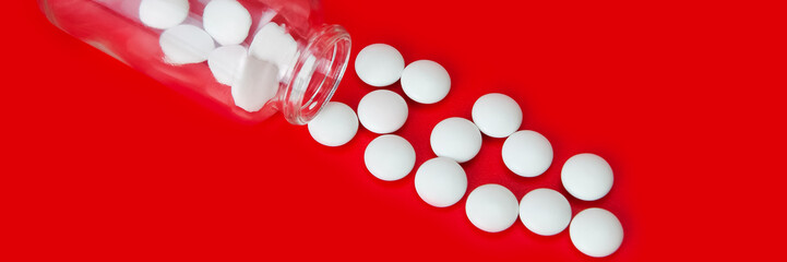 On a red background, white tablets are scattered from a glass bottle. View from above.
