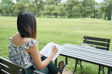 Girl wirtes something in her notebook sitting on the chair on grass field in the city park
