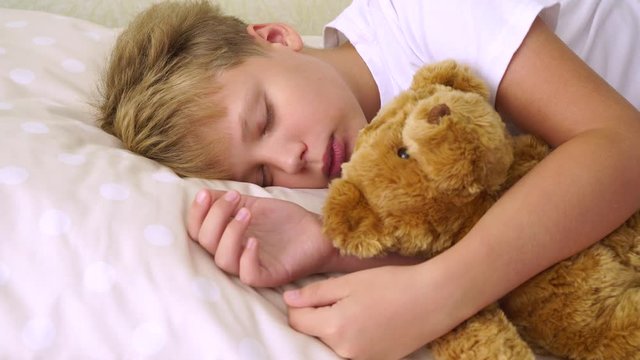Closeup portrait of cute sleepy young white kid laying in his bed with cute brown teddy bear toy. Real time 4k video footage.