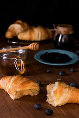 Fresh butter croissants, the glass with honey, turquoise plate with blueberry jam, blueberry jam in the glass, scattered blueberries on a dark wooden table, dark or black background.