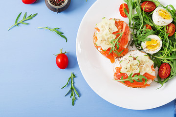 Tasty breakfast. Open sandwiches with salmon, cream cheese and rye bread in a white plate and salad with tomato, egg and arugula. Top view. Flat lay