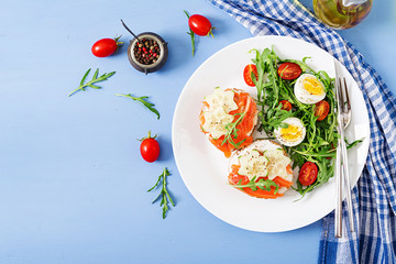Tasty breakfast. Open sandwiches with salmon, cream cheese and rye bread in a white plate and salad with tomato, egg and arugula. Top view. Flat lay