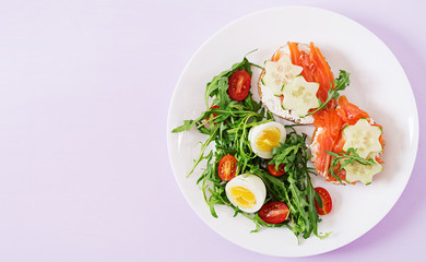Tasty breakfast. Open sandwiches with salmon, cream cheese and rye bread in a white plate and salad with tomato, egg and arugula. Top view. Flat lay