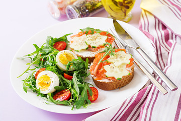 Tasty breakfast. Open sandwiches with salmon, cream cheese and rye bread in a white plate and salad with tomato, egg and arugula.