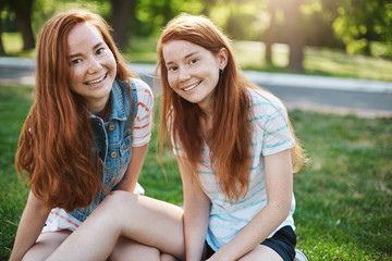 Close-up shot of beautiful european sisters with red hair and freckles sitting on green grass and smiling broadly at camera, hanging out with friends on picnic, expressing joy and amusement
