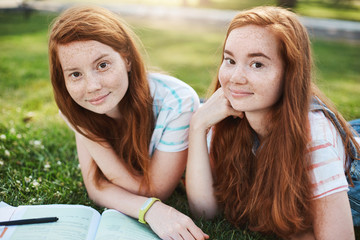 Close-up shot of two beautiful sisters with freckles and ginger hair, lying on grass, hanging out and making lessons, friend trying help girl with homework, spending time with joy and pleasure