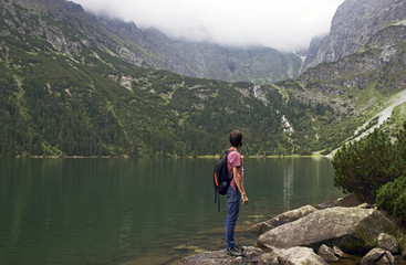 A young man with a backpack stands against the background of mountains and lake. rear view. the guy looks at the beautiful view of nature. sunny summer day. Dombay. Russia.