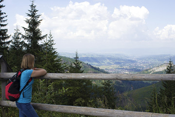 Fototapeta na wymiar a girl in a blue T-shirt with a red briefcase leaned on the railing and looked at the mountains, at home and in the sky. road of a tourist to the mountain