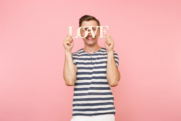 Portrait of emotional young man in striped t-shirt hold wooden word Love isolated on trending pastel pink background. People sincere emotions lifestyle holiday concept. Advertising area for copy space