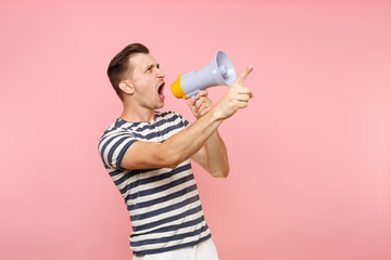 Portrait of angry young man in striped t-shirt swearing in electronic megaphone on copy space isolated on trending pastel pink background. People sincere emotions lifestyle concept. Advertising area.