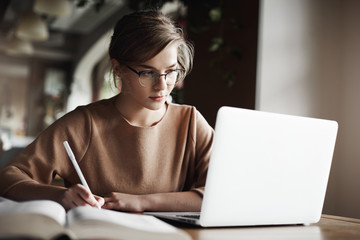 Creative good-looking european female with fair hair in trendy glasses, making notes while looking at laptop screen, working or preparing for business meeting, being focused and hardworking
