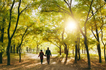 Back rear view of young couple in love woman with backpack, man in casual warm clothes walking by hands on road in autumn city park outdoors in sunny day. Love relationship family lifestyle concept.