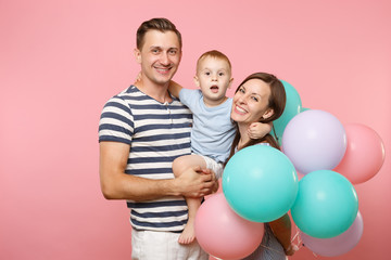 Fototapeta na wymiar Portrait of young happy family, parents keep in arms, kissing hugging child kid son baby boy, celebrating birthday holiday party on pink background with colorful air balloons. Sincere emotions concept