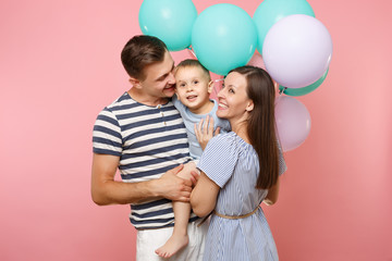Portrait of young happy family, parents keep in arms, kissing hugging child kid son baby boy, celebrating birthday holiday party on pink background with colorful air balloons. Sincere emotions concept