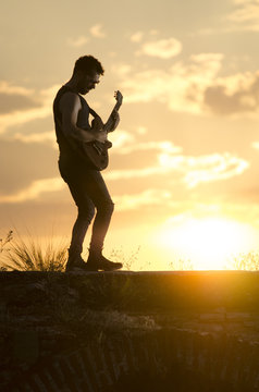 Man performs on electric guitar on old Roman aqueduct during sunset.