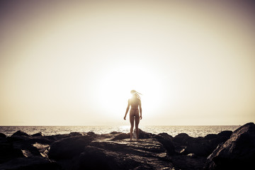 standing back rear view portrait for fitness body young female enjoying the golden dark sunset at the rocky beach. horizon and quiet ocean in background. wind in the hair. summer vacation ending conce