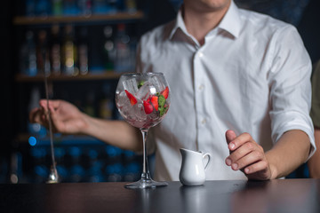 Wineglass filled with ice, strawberries and white milk pitcher is on the bar counter. Process of a cocktail preparation.