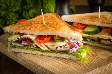 Close-up of two sandwiches with bacon, salami, prosciutto and fresh vegetables on rustic wooden cutting board. Club sandwich concept.