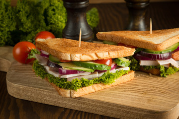 Close-up of two sandwiches with bacon, salami, prosciutto and fresh vegetables on rustic wooden cutting board. Club sandwich concept.