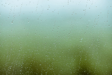 Dirty glass with drops of rain. Raindrops on green and blue clear background. Greenery and sky in bokeh outside window. Droplets and stains close up. Detailed transparent texture in macro.