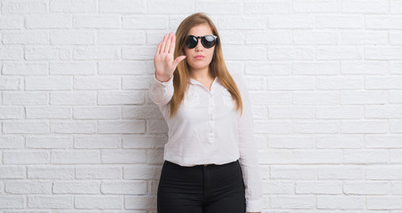 Young adult business woman standing over white brick wall wearing sunglasses with open hand doing...