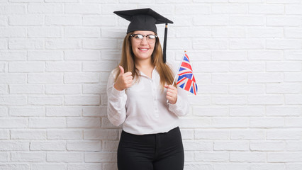 Young adult woman over white brick wall wearing graduate cap holding united kingdom flag happy with...