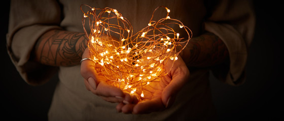 Bright garlands in the shape of a heart, holding female hands with a tattoo on a dark background.