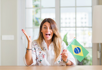 Young woman at home holding flag of Brazil very happy and excited, winner expression celebrating victory screaming with big smile and raised hands