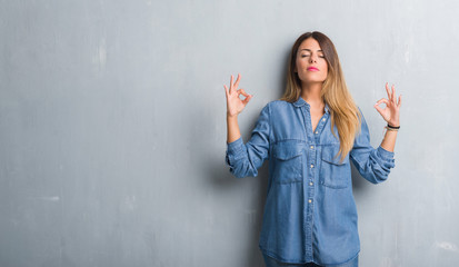 Young adult woman over grunge grey wall wearing denim outfit relax and smiling with eyes closed doing meditation gesture with fingers. Yoga concept.