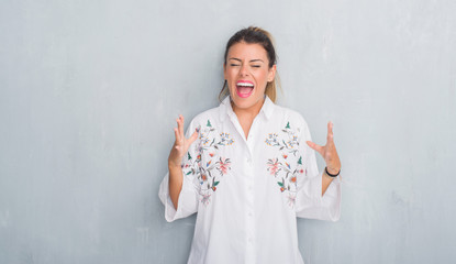 Young adult woman over grunge grey wall wearing flowers shirt celebrating mad and crazy for success with arms raised and closed eyes screaming excited. Winner concept