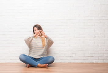 Young adult woman sitting on the floor over white brick wall at home smiling in love showing heart symbol and shape with hands. Romantic concept.