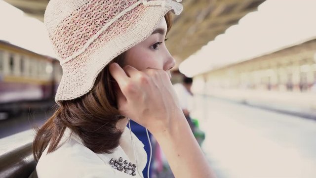 Happy teen Asian woman listening to the music with headphones and waiting in a train station summer. Travel Thailand concept.