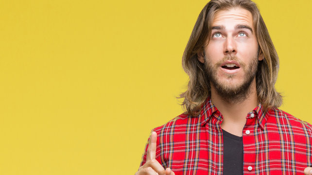 Young handsome man with long hair over isolated background amazed and surprised looking up and pointing with fingers and raised arms.
