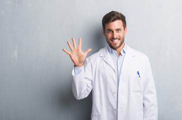 Handsome young professional man over grey grunge wall wearing white coat showing and pointing up with fingers number five while smiling confident and happy.