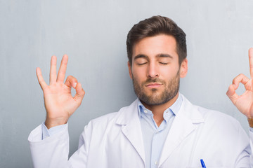 Handsome young professional man over grey grunge wall wearing white coat relax and smiling with eyes closed doing meditation gesture with fingers. Yoga concept.