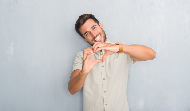 Handsome young man over grey grunge wall wearing summer shirt smiling in love showing heart symbol and shape with hands. Romantic concept.