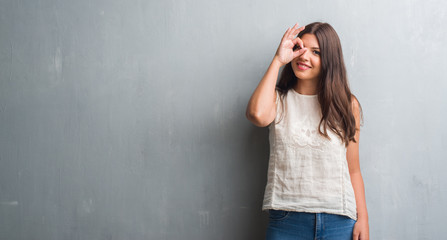 Young brunette woman over grunge grey wall with happy face smiling doing ok sign with hand on eye looking through fingers