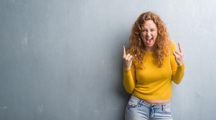 Young redhead woman over grey grunge wall shouting with crazy expression doing rock symbol with hands up. Music star. Heavy concept. - obrazy, fototapety, plakaty