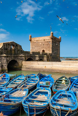 Eine Flotte blauer Fischerboote drängt sich im Hafen von Essaouira in Marokko zusammen. Sie können auch die Befestigungsanlagen und einen Turm der Zitadelle von Mogador sehen