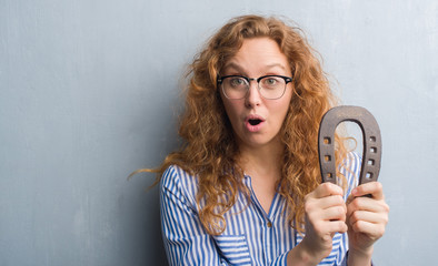 Young redhead woman over grey grunge wall holding horseshoe scared in shock with a surprise face,...