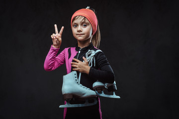Confident little skater girl dressed in sportswear holds ice skates and shows victory hand. Isolated on a dark textured background.