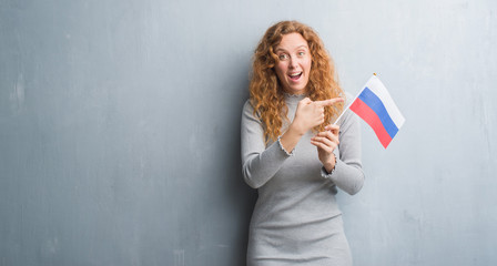 Young redhead woman over grey grunge wall holding flag of Russia very happy pointing with hand and finger
