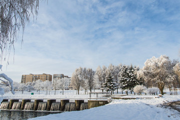 winter park, different trees covered with snow, artificial waterfall on the background of houses