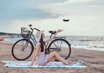 Fototapeta premium Portrait of a young brunette girl in sunglasses and hat wearing dress enjoy the vacation on the beach, playing with a quadcopter.