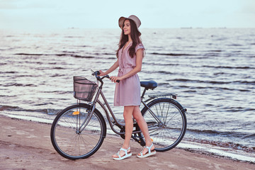 Fototapeta premium Happy sensual girl in hat wearing dress walks with her bicycle on the beach against amazing seaside background.