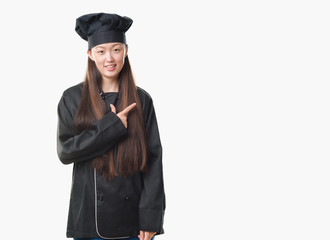 Young Chinese woman over isolated background wearing chef uniform cheerful with a smile of face pointing with hand and finger up to the side with happy and natural expression on face