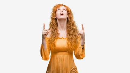 Young redhead woman amazed and surprised looking up and pointing with fingers and raised arms.