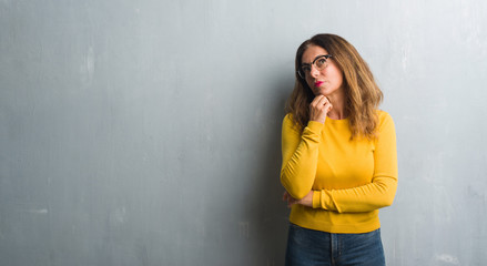 Fototapeta premium Middle age hispanic woman over grey wall wearing glasses with hand on chin thinking about question, pensive expression. Smiling with thoughtful face. Doubt concept.